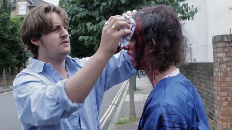 A man in a blue shirt wipes blood from another man’s head in a suburban street