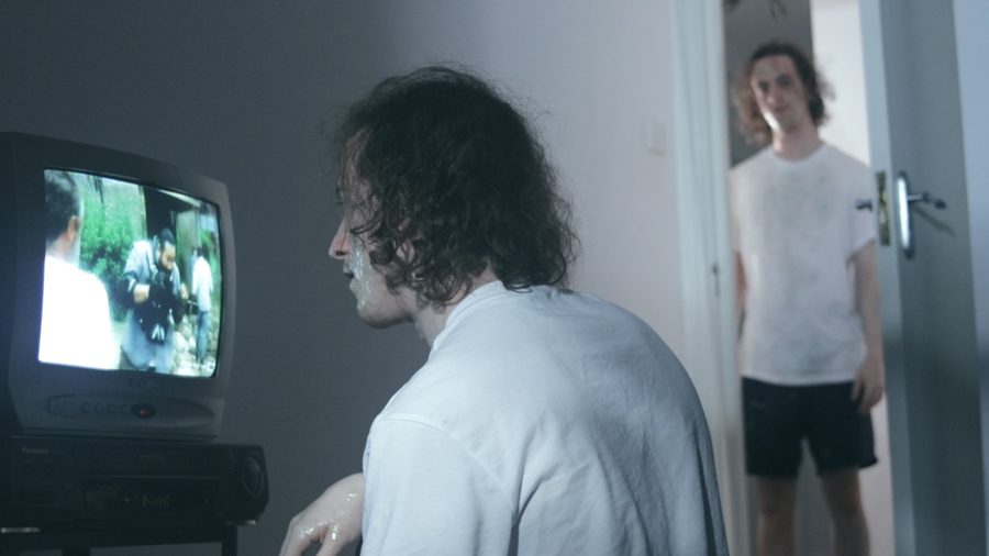 A man watches an old CRT television while another stands in the doorway