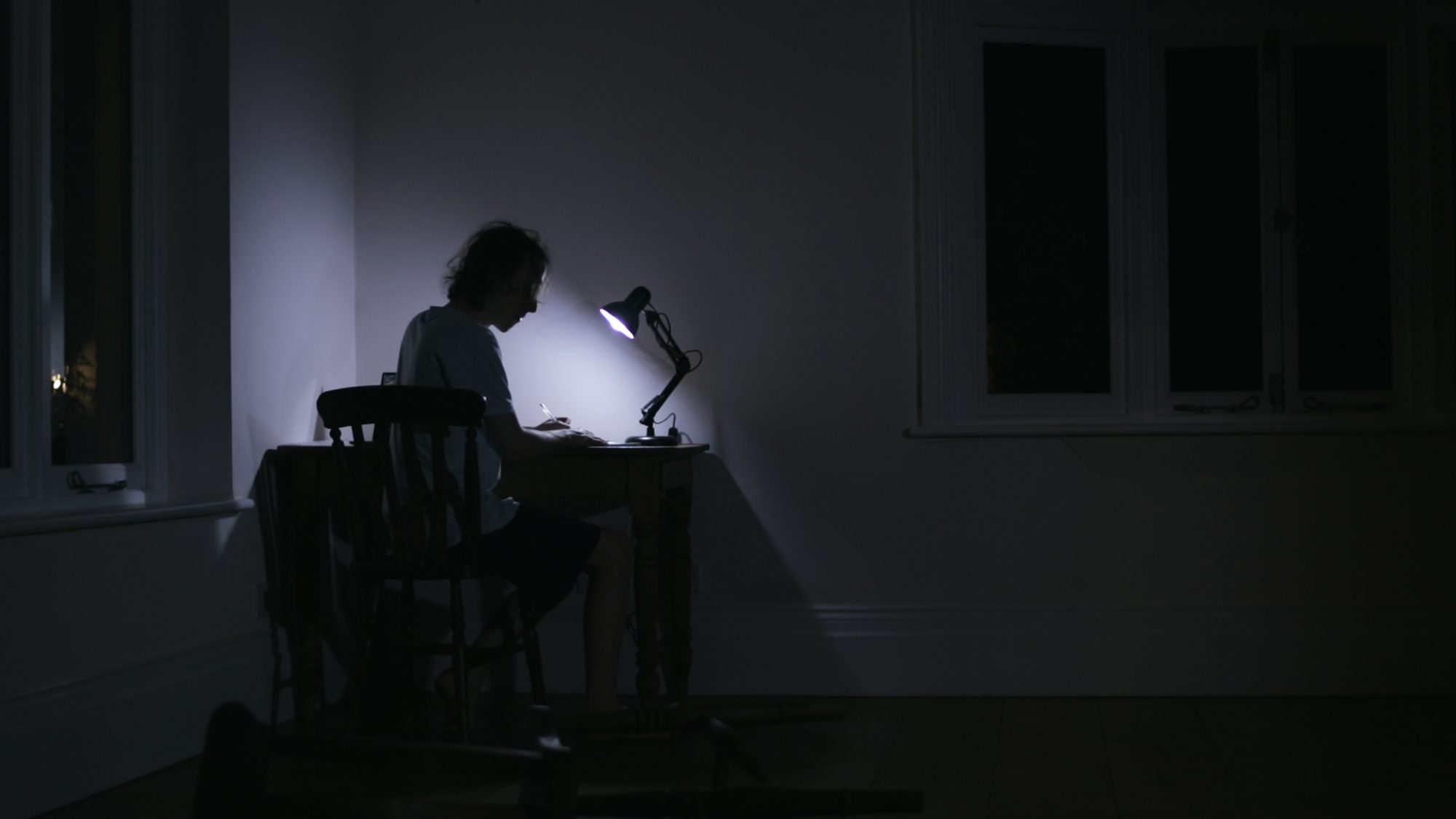 A man works alone at a desk under a single lamp in a dark room