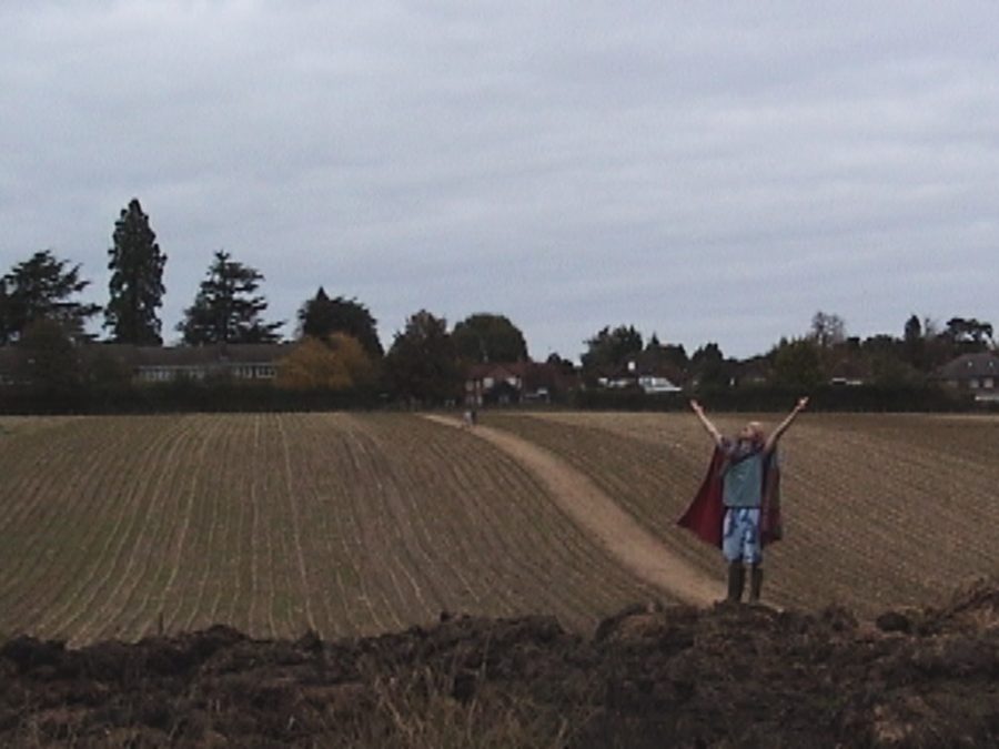 A figure in a cape raises both arms in a ploughed field